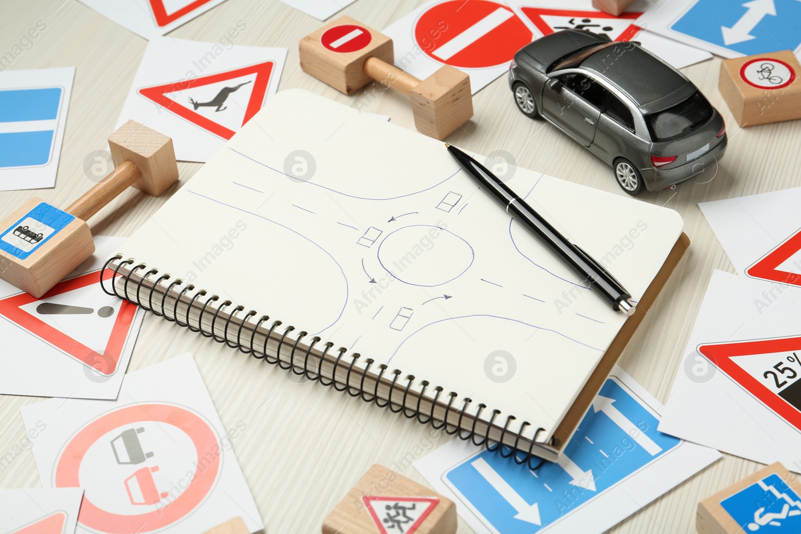 Photo of Many different road signs, notebook with sketch of roundabout and toy car on white wooden background. Driving school