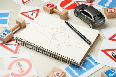 Photo of Many different road signs, notebook with sketch of roundabout and toy car on white wooden background. Driving school