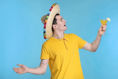 Young man in Mexican sombrero hat with cocktail on light blue background