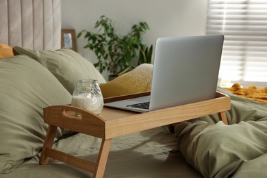 Photo of Wooden tray with modern laptop and burning candle on bed indoors