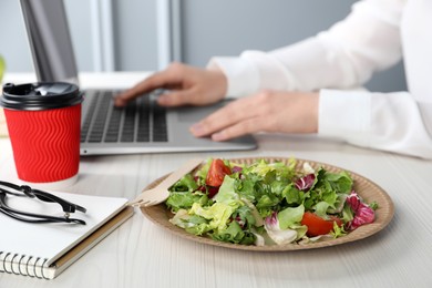 Photo of Office employee working with laptop at white wooden table, focus on vegetable salad and paper cup of coffee. Business lunch