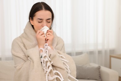 Sick woman wrapped in blanket with tissue blowing nose at home, space for text. Cold symptoms