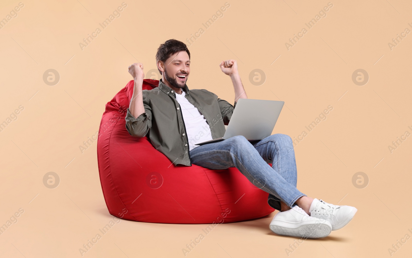 Photo of Happy man with laptop sitting in beanbag chair against beige background