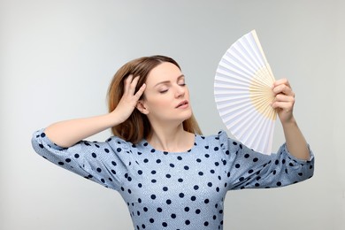Beautiful woman waving hand fan to cool herself on light grey background