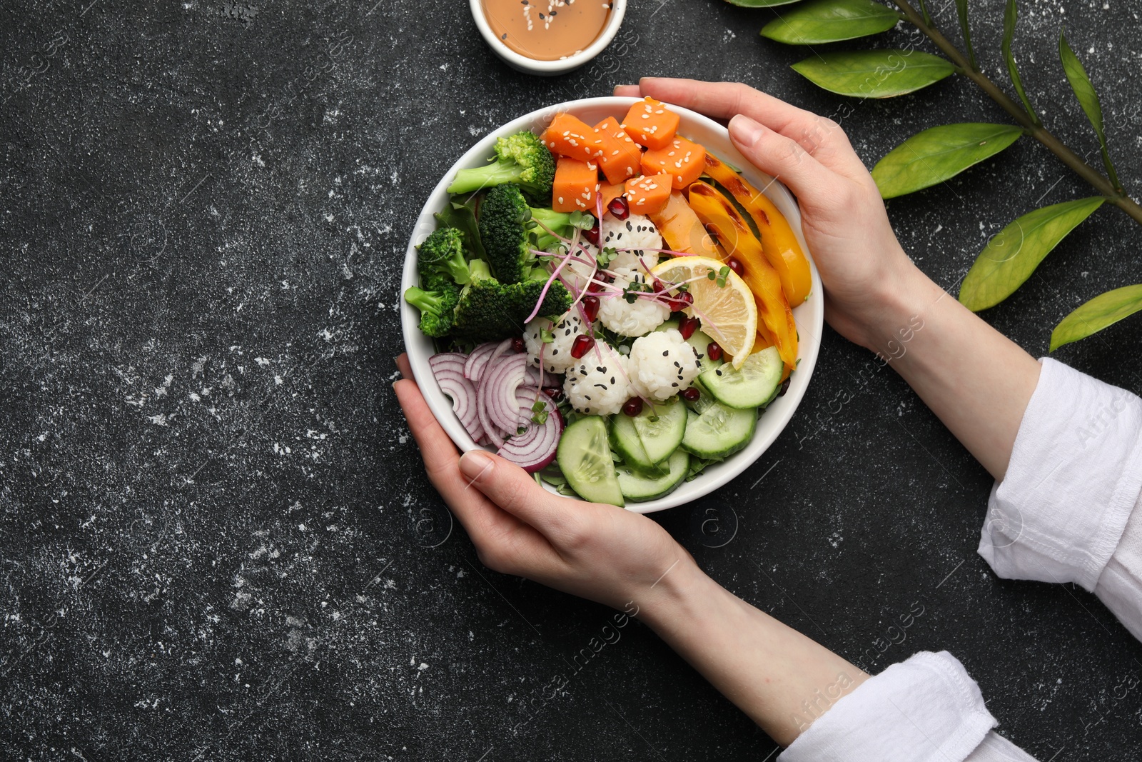 Photo of Woman holding bowl with many different vegetables and rice at grey textured table, top view and space for text. Vegan diet