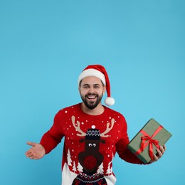 Photo of Happy young man in Christmas sweater and Santa hat holding gift on light blue background