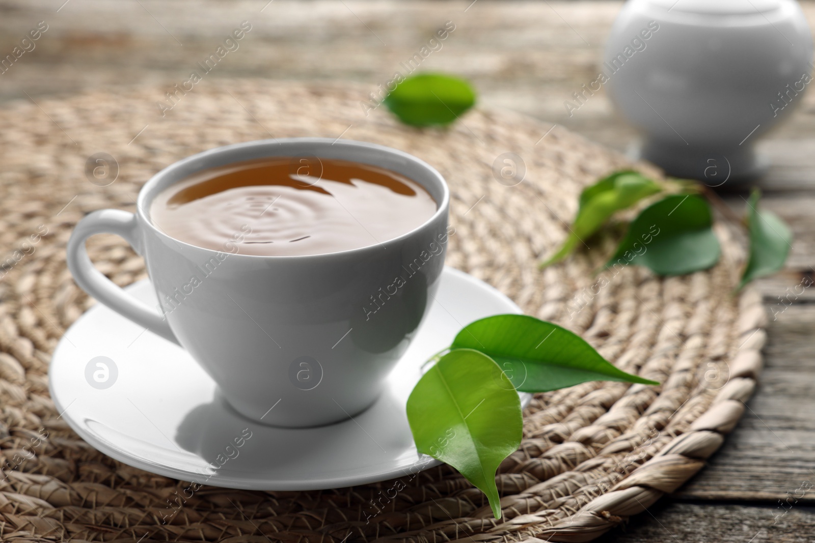 Photo of Green tea in white cup with leaves and wicker mat on table, closeup