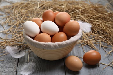 Photo of Fresh chicken eggs in bowl, feathers and dried straw on grey wooden table