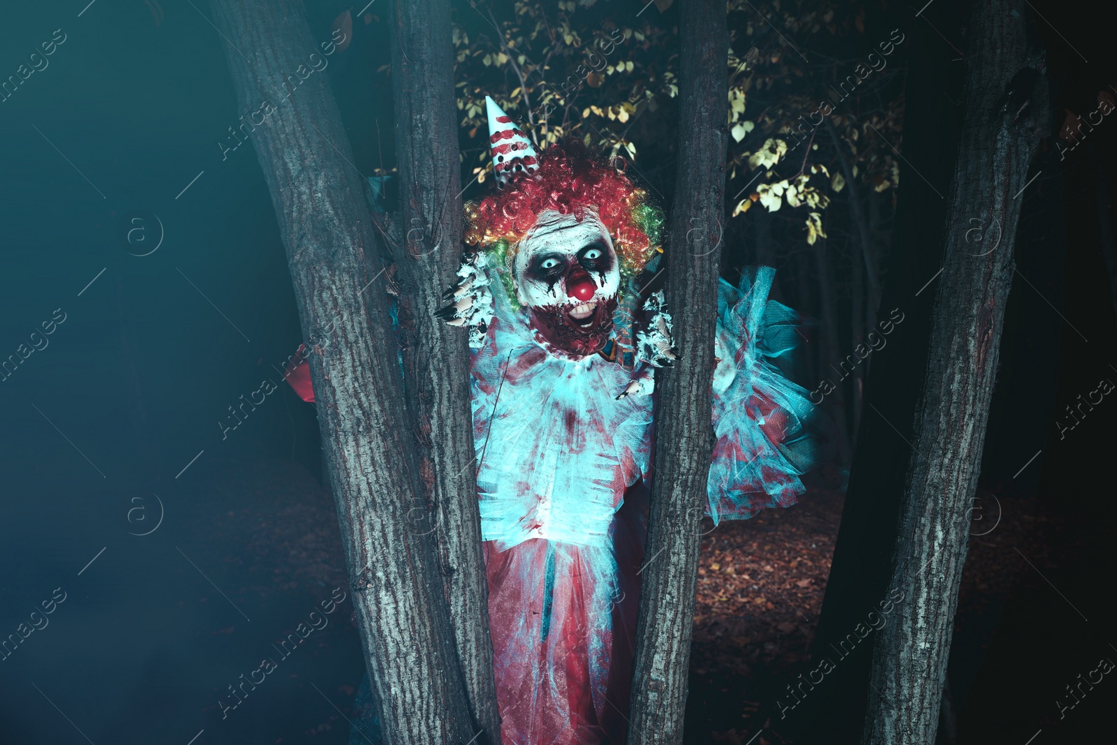 Photo of Terrifying clown hiding behind trees outdoors at night. Halloween party costume