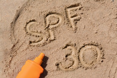 Photo of Abbreviation SPF 30 written on sand and bottle of sunscreen at beach, top view