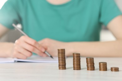 Financial savings. Woman making notes at white wooden table, focus on stacked coins