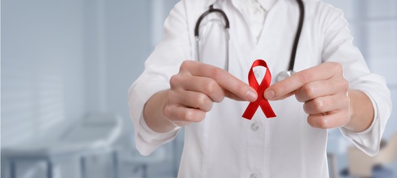 Cancer awareness. Doctor holding red ribbon on blurred background, closeup. Banner design with space for text