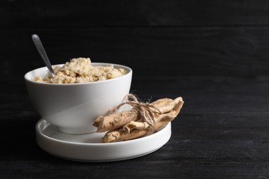 Photo of Bowl of tasty prepared horseradish, spoon and roots on black wooden table. Space for text