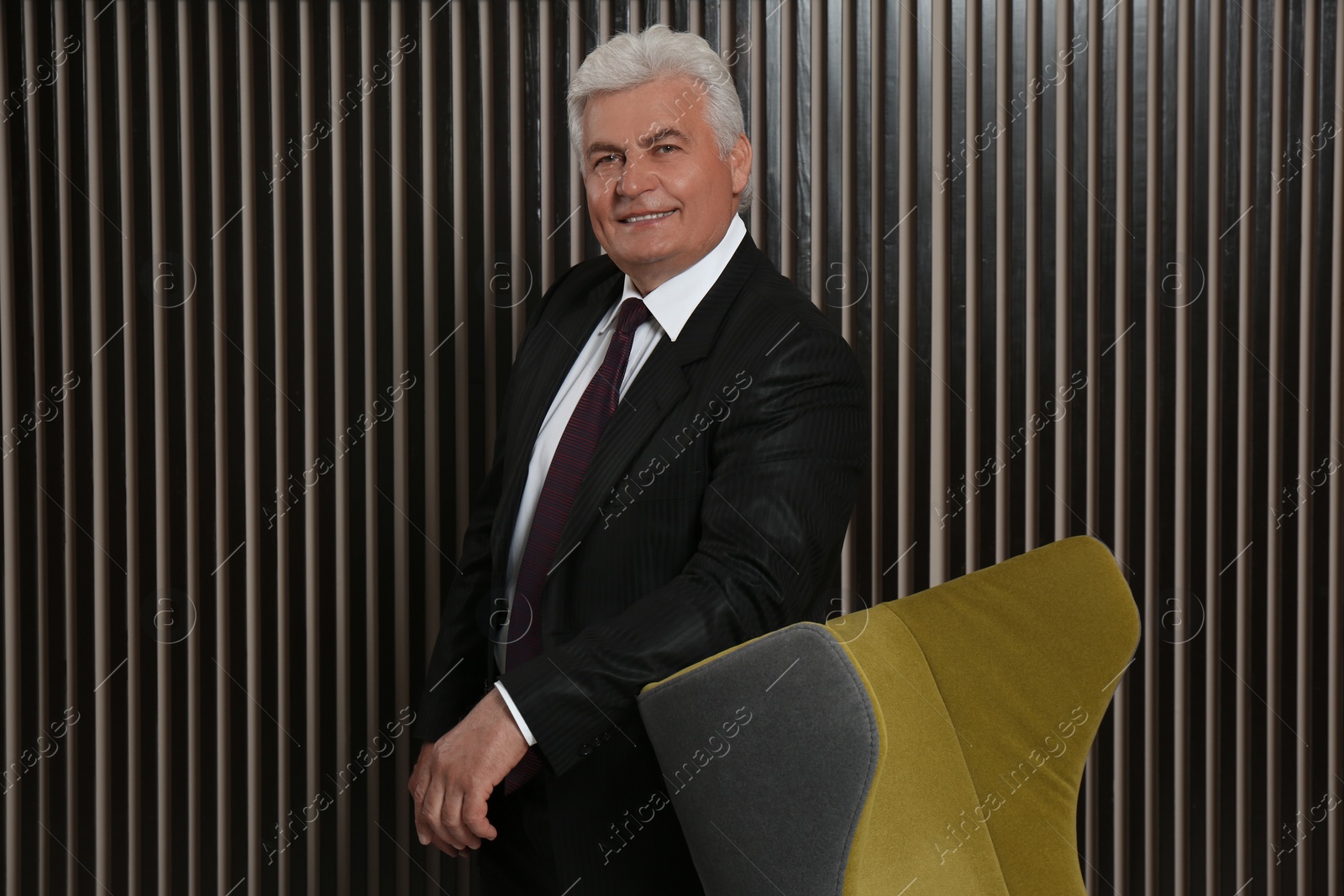 Photo of Happy mature businessman standing near armchair  near wood slat wall