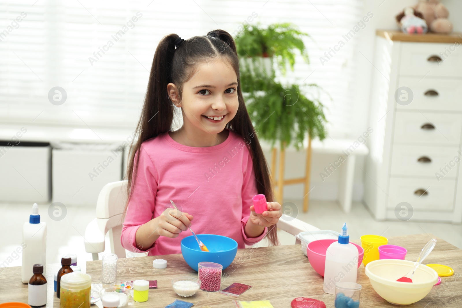 Photo of Cute little girl mixing ingredients with silicone spatula at table. DIY slime toy