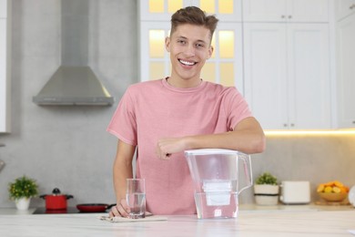 Happy man with filter jug and glass of clear water at table in kitchen