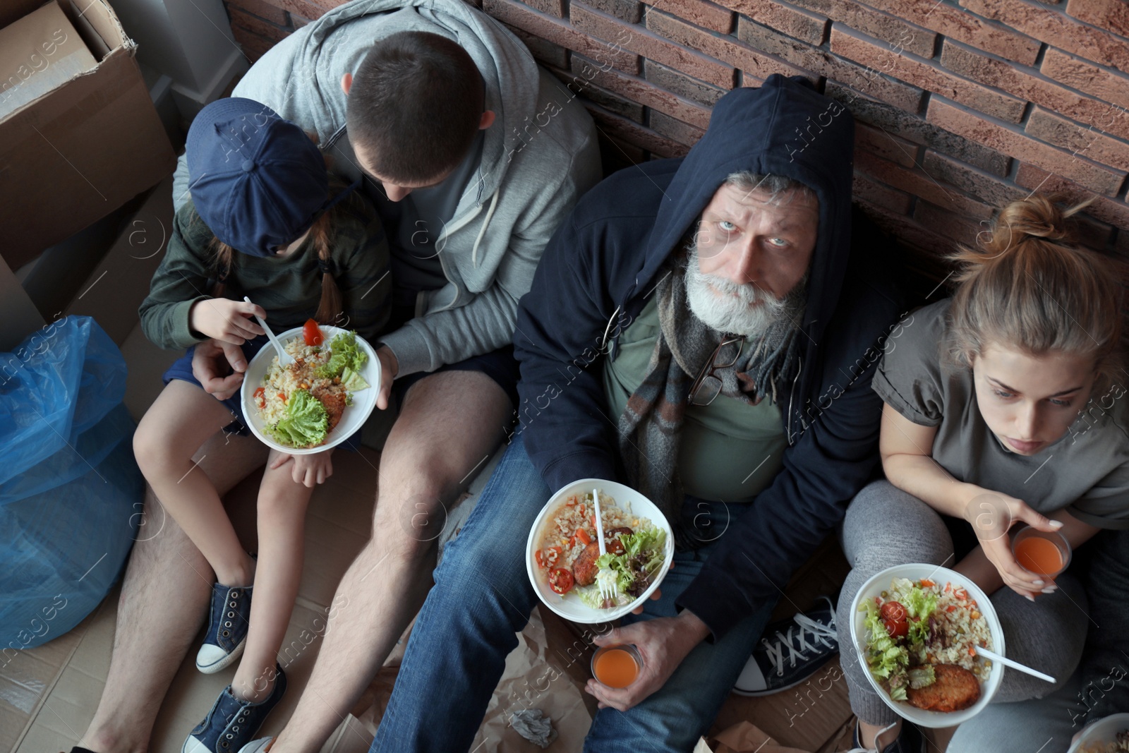 Photo of Poor people with plates of food sitting at wall indoors, view from above