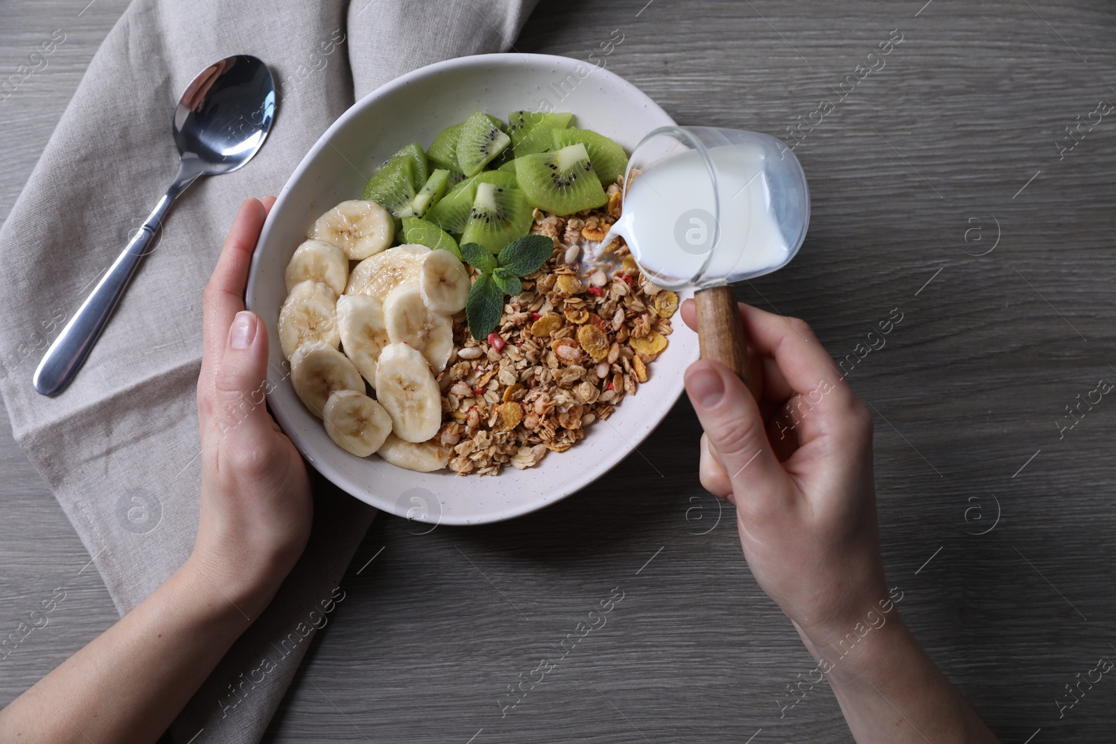 Photo of Woman pouring milk into bowl of tasty granola with banana and kiwi at grey wooden table, top view