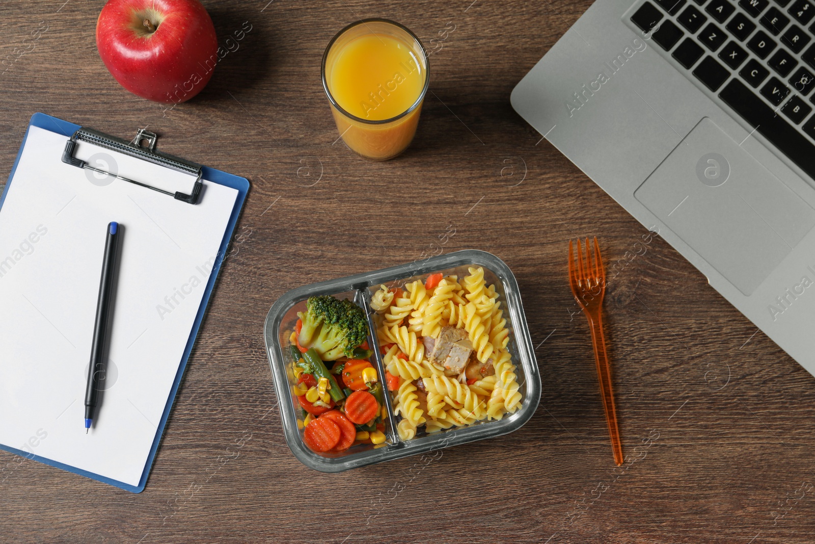 Photo of Container of tasty food, fork, laptop, glass of juice, apple and clipboard on wooden table, flat lay. Business lunch