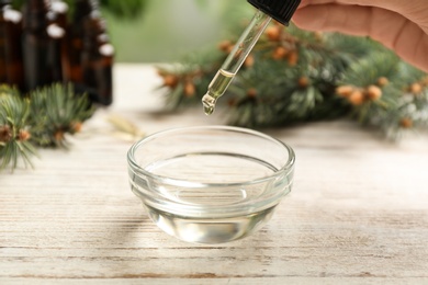 Woman holding pipette with conifer essential oil over bowl on wooden table, closeup