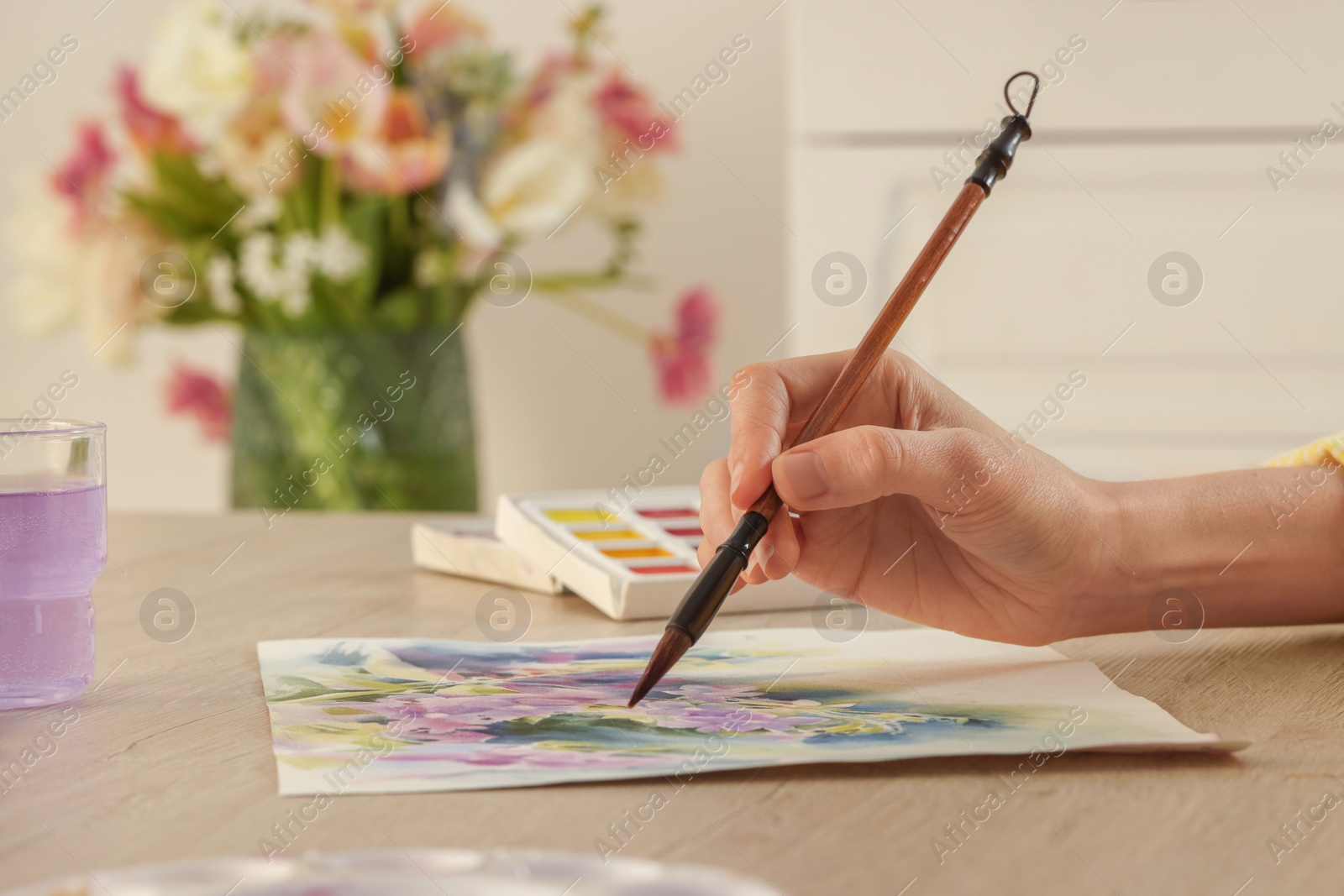 Photo of Woman painting flowers with watercolor at white wooden table indoors, closeup. Creative artwork