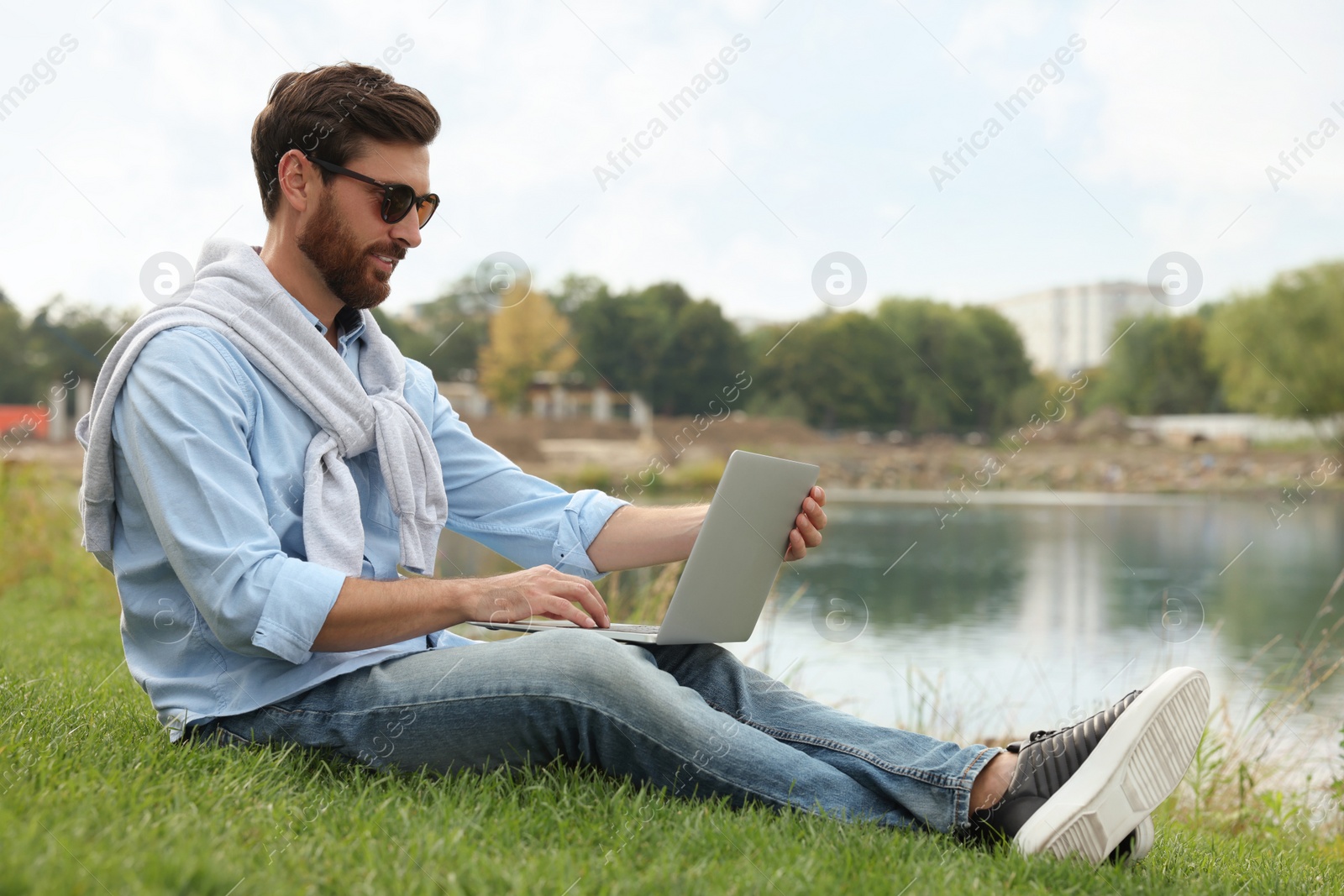 Photo of Man in sunglasses with laptop on green grass near lake