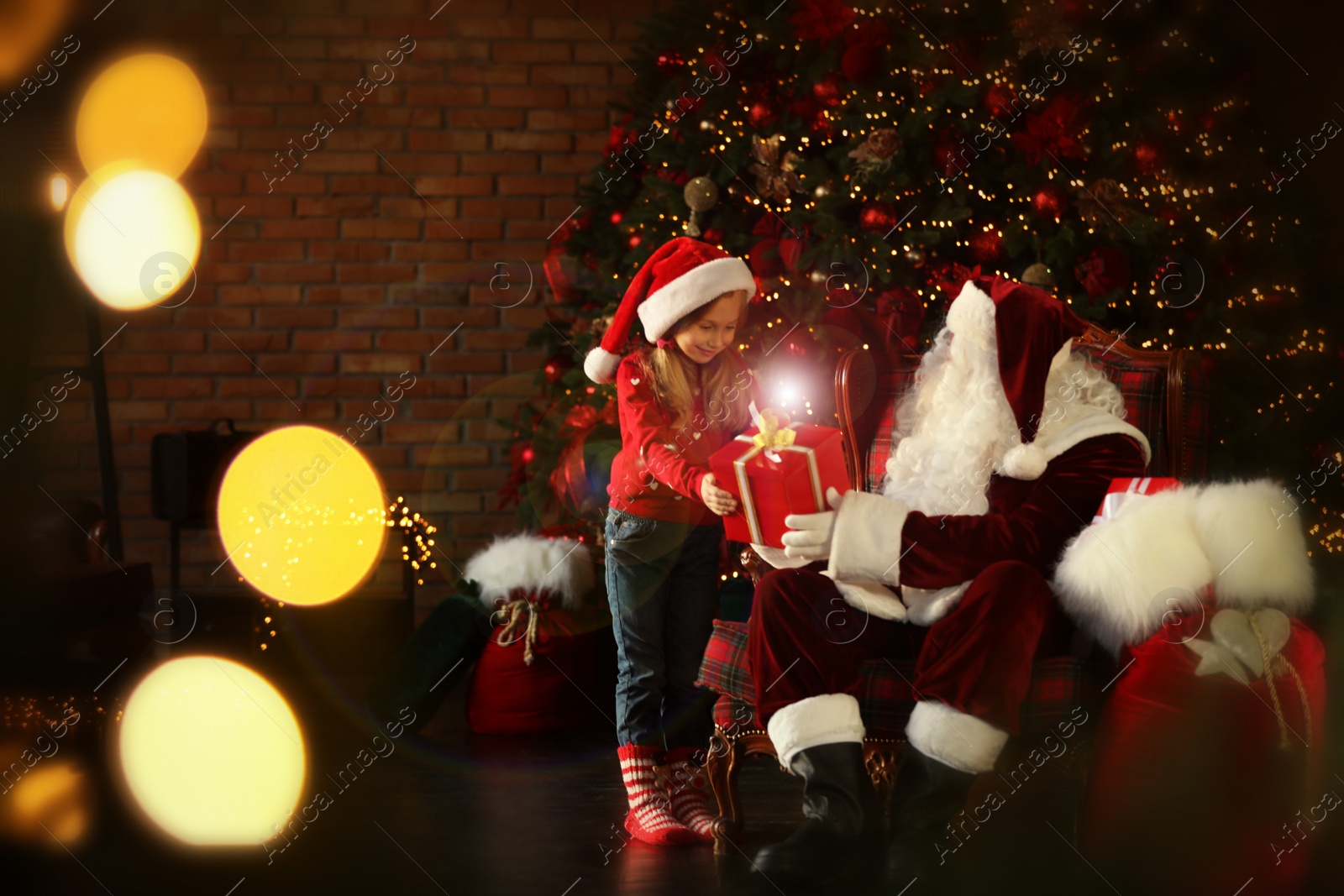 Photo of Santa Claus giving present to little girl near Christmas tree indoors