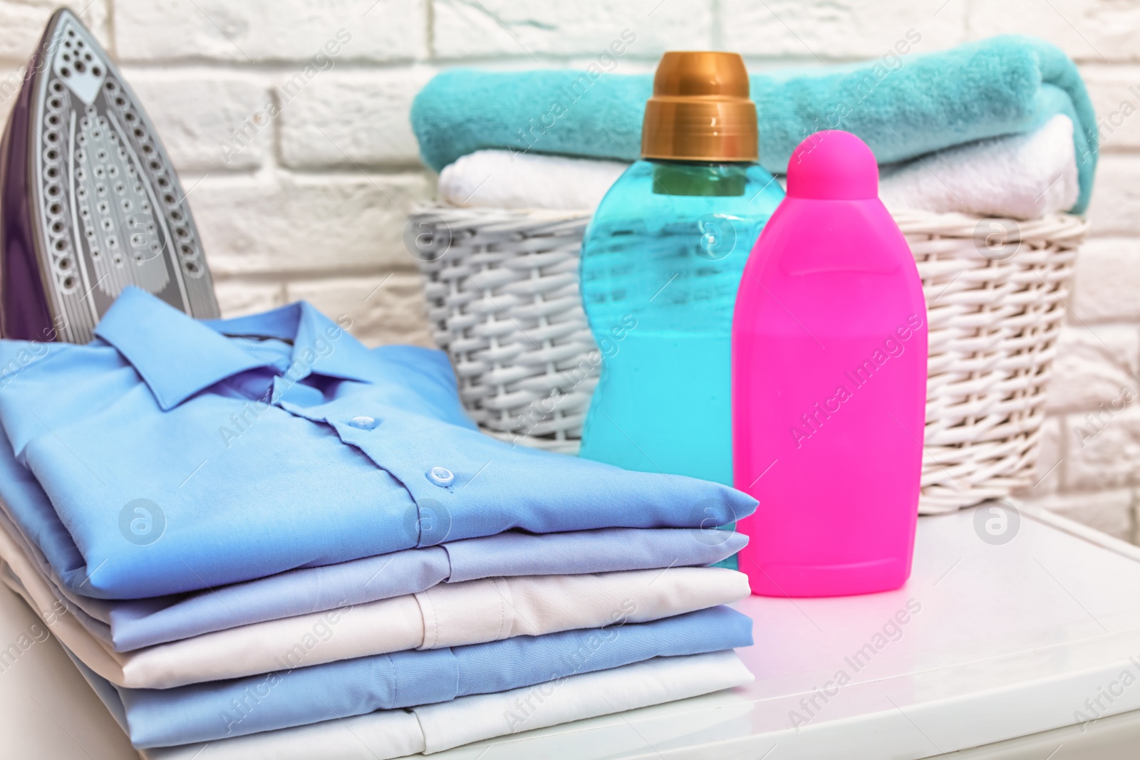 Photo of Stack of clean clothes, iron, detergents and basket with towels on table