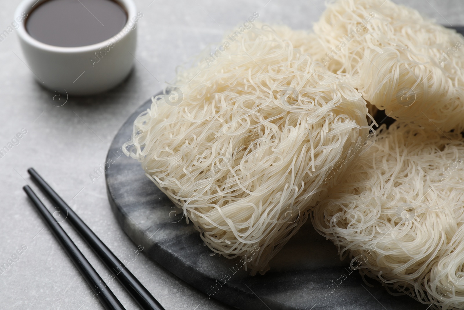 Photo of Uncooked rice noodles, soy sauce and chopsticks on light grey table, closeup