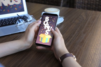Image of Bonus gaining. Woman holding smartphone on screen at wooden table, closeup. Illustration of open gift box, word and confetti on device screen