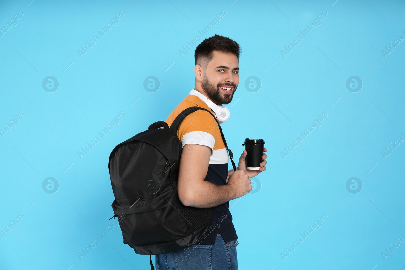 Photo of Young man with stylish backpack, headphones and cup of coffee on light blue background