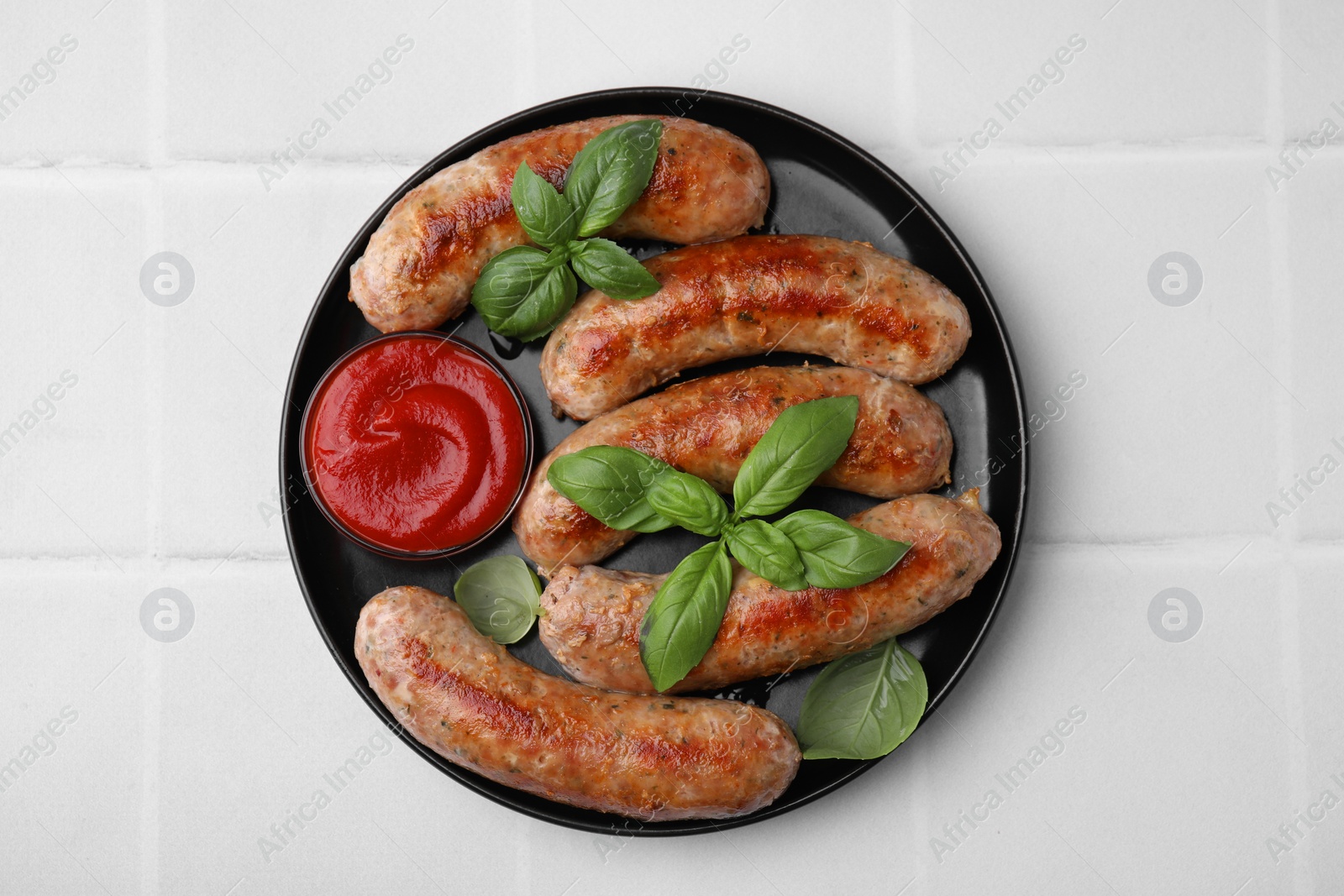 Photo of Plate with tasty homemade sausages, ketchup and basil leaves on white tiled table, top view