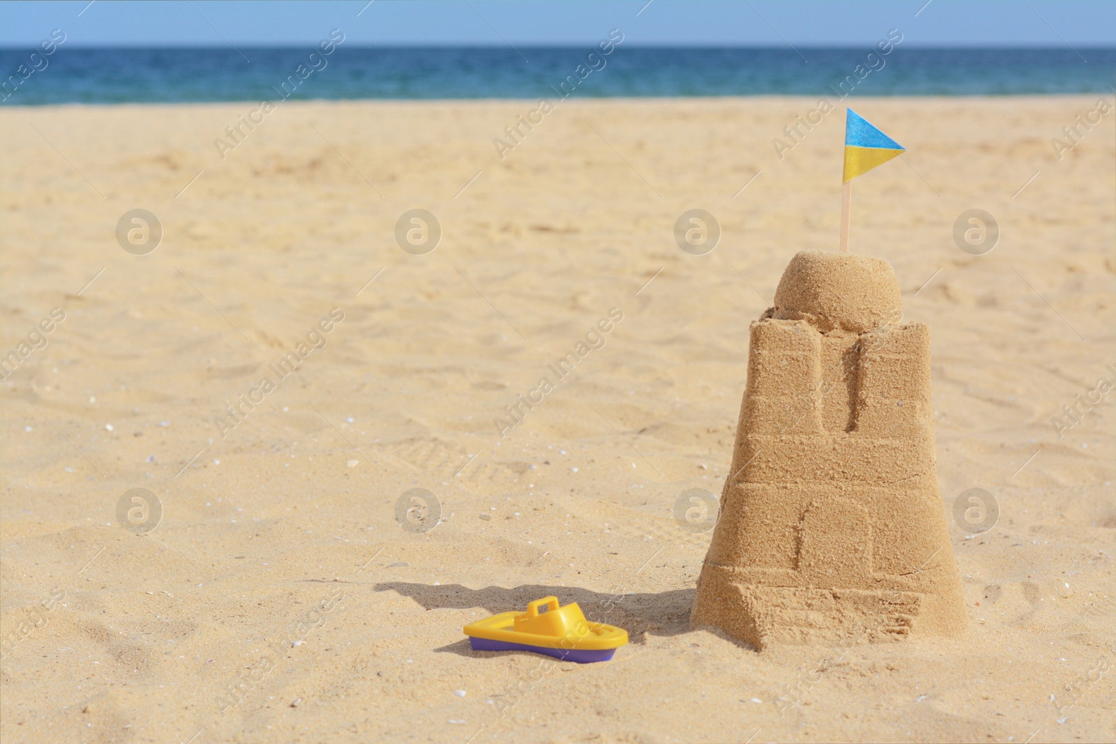 Photo of Beautiful sand castle with small Ukrainian flag and toy boat on beach near sea, space for text