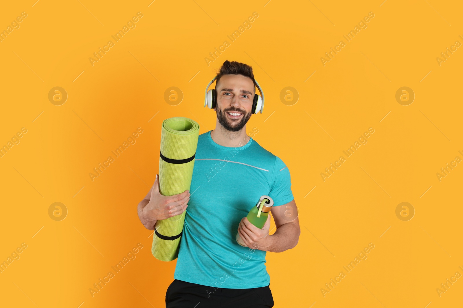 Photo of Handsome man with yoga mat, headphones and bottle on yellow background