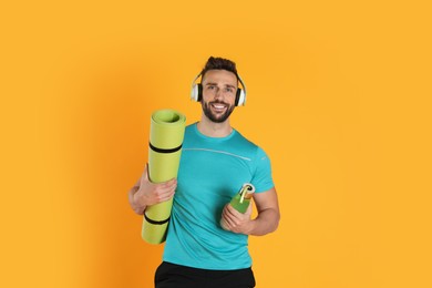 Handsome man with yoga mat, headphones and bottle on yellow background