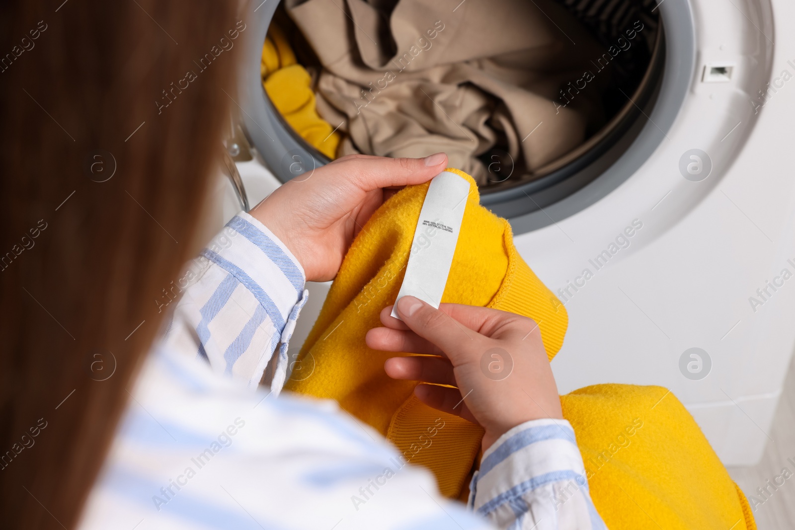 Photo of Woman reading clothing label with care symbols and material content on yellow shirt near washing machine, closeup