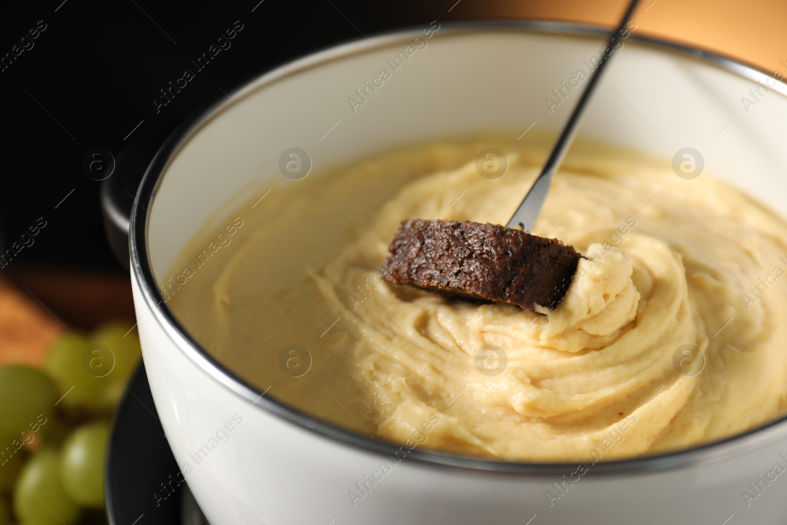 Photo of Dipping piece of bread into fondue pot with melted cheese on dark background, closeup