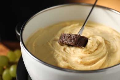 Dipping piece of bread into fondue pot with melted cheese on dark background, closeup