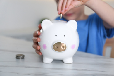 Photo of Little boy putting coin into piggy bank at marble table indoors, closeup