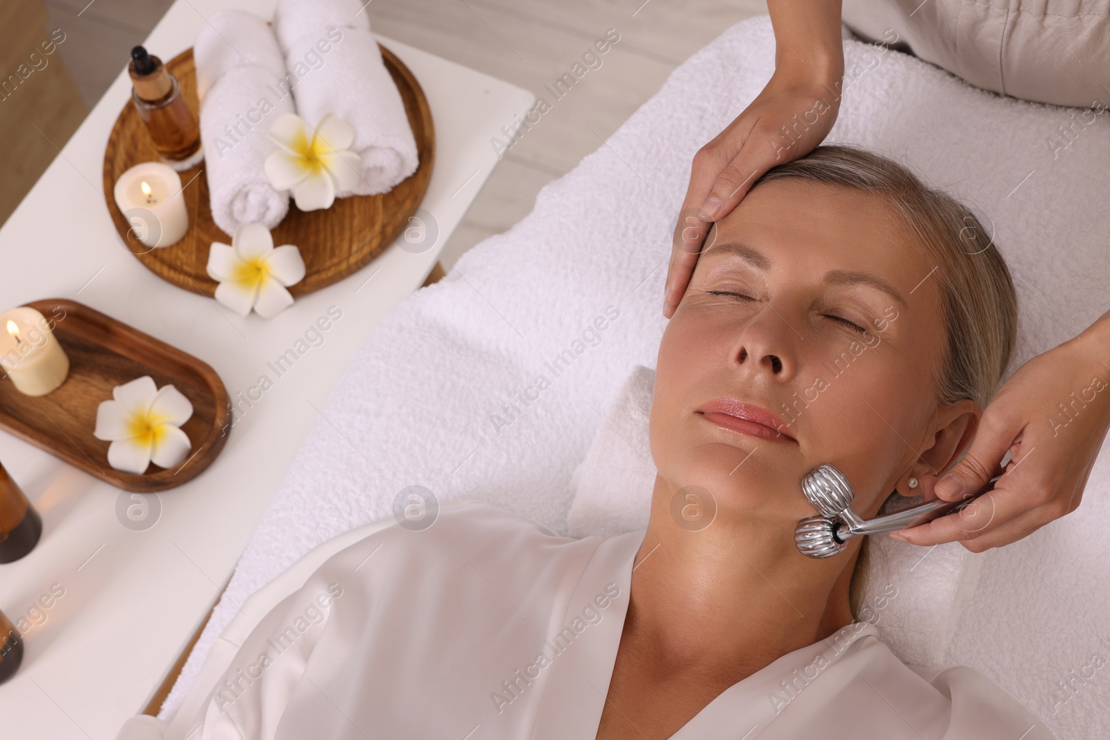 Photo of Woman receiving facial massage with metal roller in beauty salon, above view