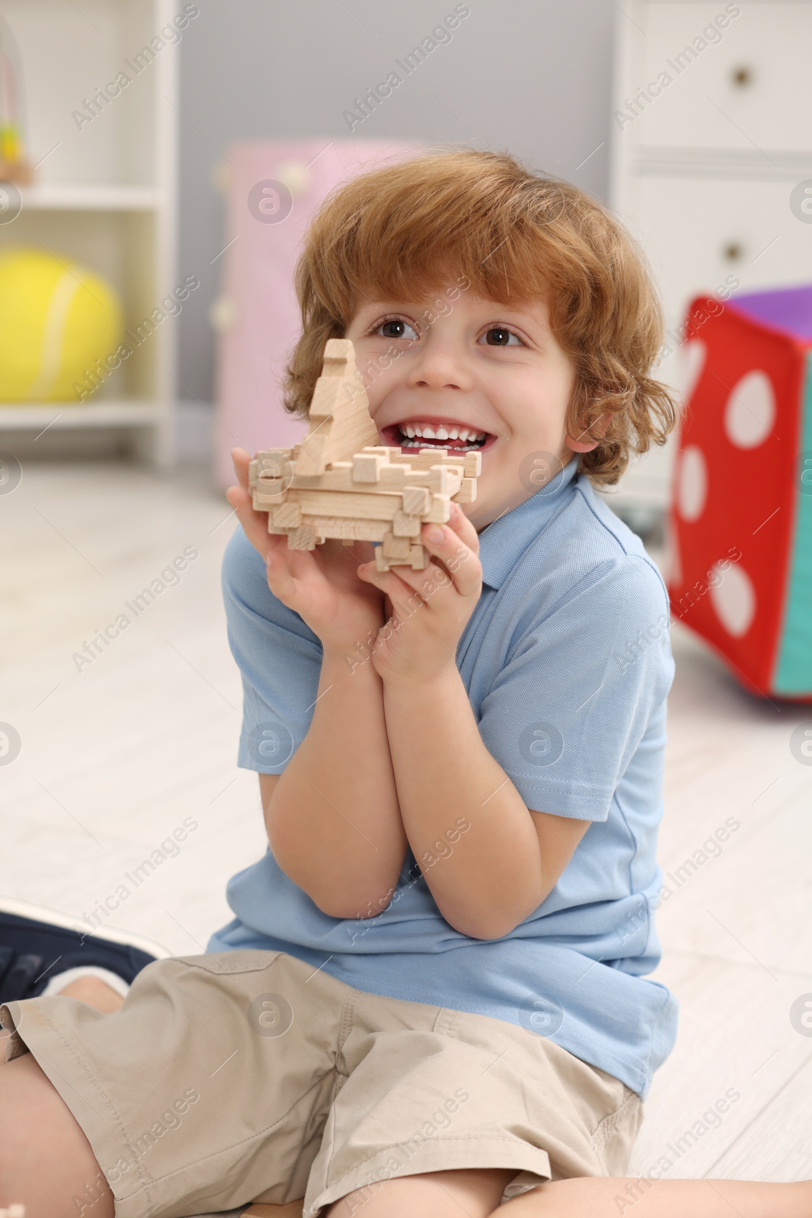 Photo of Happy little boy playing with piece of wooden construction set on floor in room. Child's toy