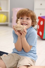 Photo of Happy little boy playing with piece of wooden construction set on floor in room. Child's toy