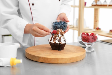 Photo of Female pastry chef decorating cupcake with berries at table in kitchen, closeup