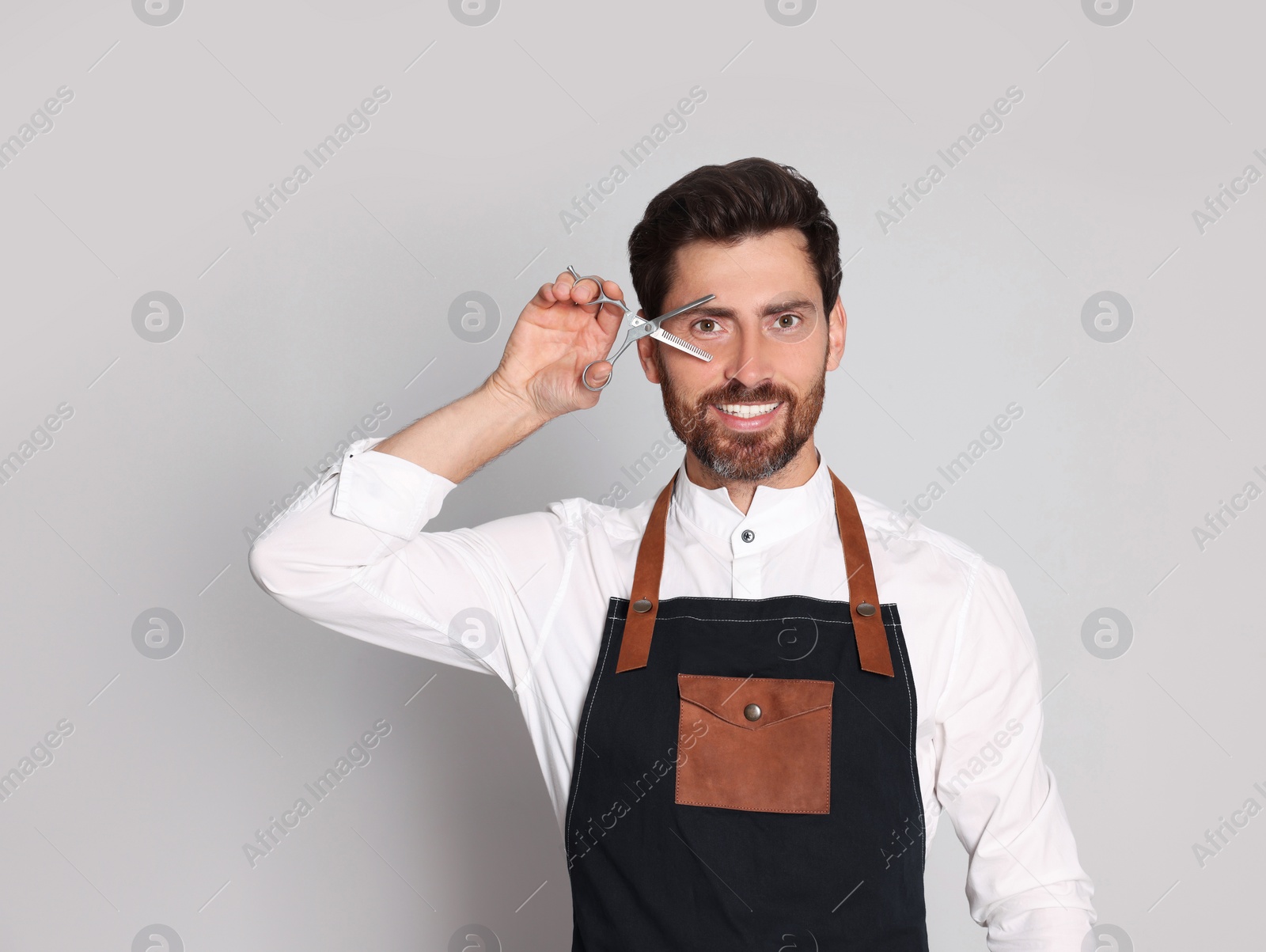 Photo of Smiling hairdresser wearing apron holding scissors on light grey background