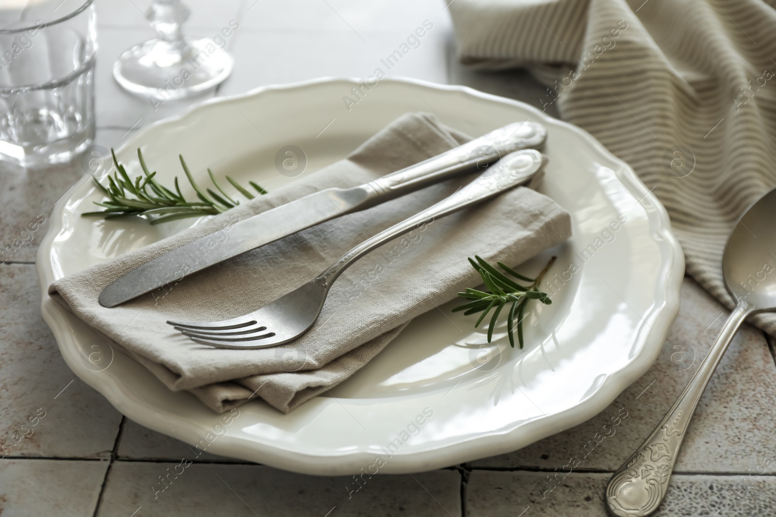 Photo of Stylish setting with cutlery, napkin, rosemary and plate on light tiled table, closeup