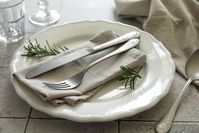 Stylish setting with cutlery, napkin, rosemary and plate on light tiled table, closeup