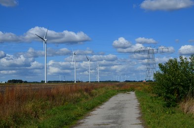 Pathway, high voltage tower with electricity transmission power lines and windmills in field on sunny day