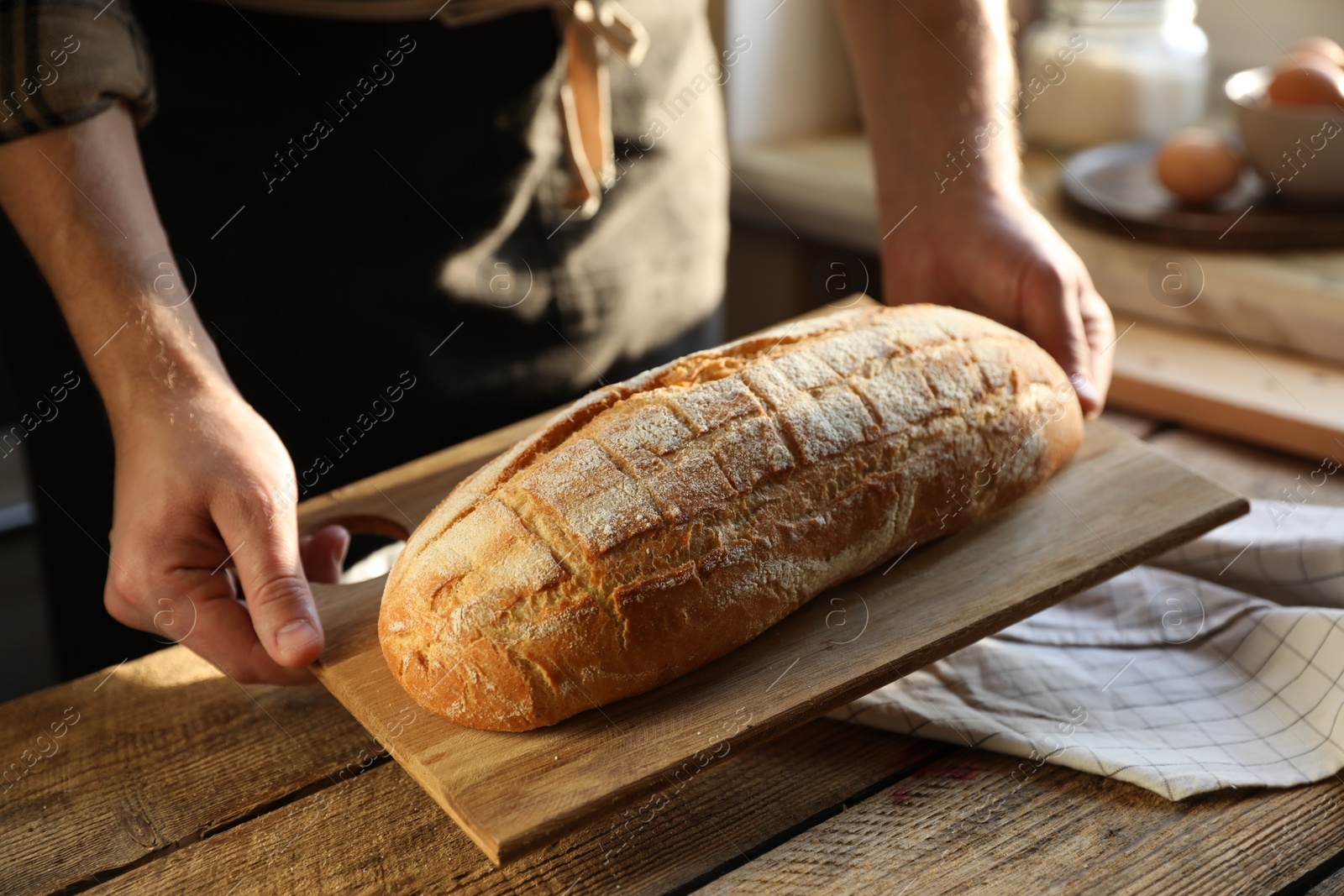 Photo of Man holding loaf of fresh bread at wooden table indoors, closeup