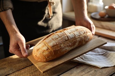 Photo of Man holding loaf of fresh bread at wooden table indoors, closeup