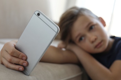 Photo of Sad little child with smartphone leaning on sofa in room. Danger of internet